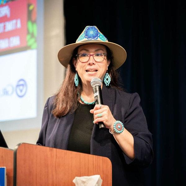 Martha Villegas Miranda at a podium with microphone, wearing a hat