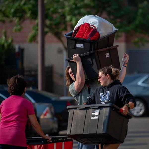 A person holds a plastic bin while another standing behind the person hoists two plastic bins of belongings.