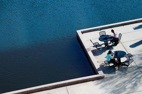  从上面看，学生们坐在桌子旁，沿着蓝色的池塘享用午餐. 